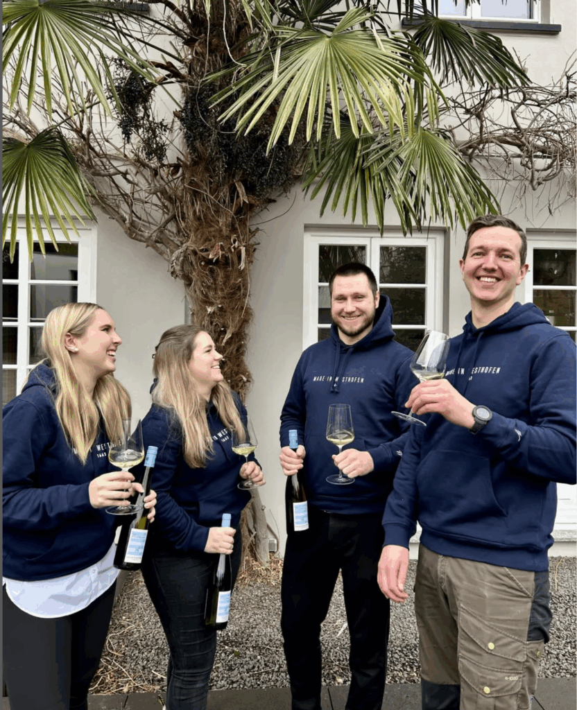 Two women and two men smile and laugh while holding bottles and glasses of white wine, taken in front of the Wittmann estate.
